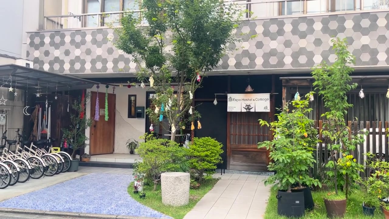Entrance of Expo Hostel in Nishijin, Kyoto, with a garden tree, potted plants and a wooden lattice door