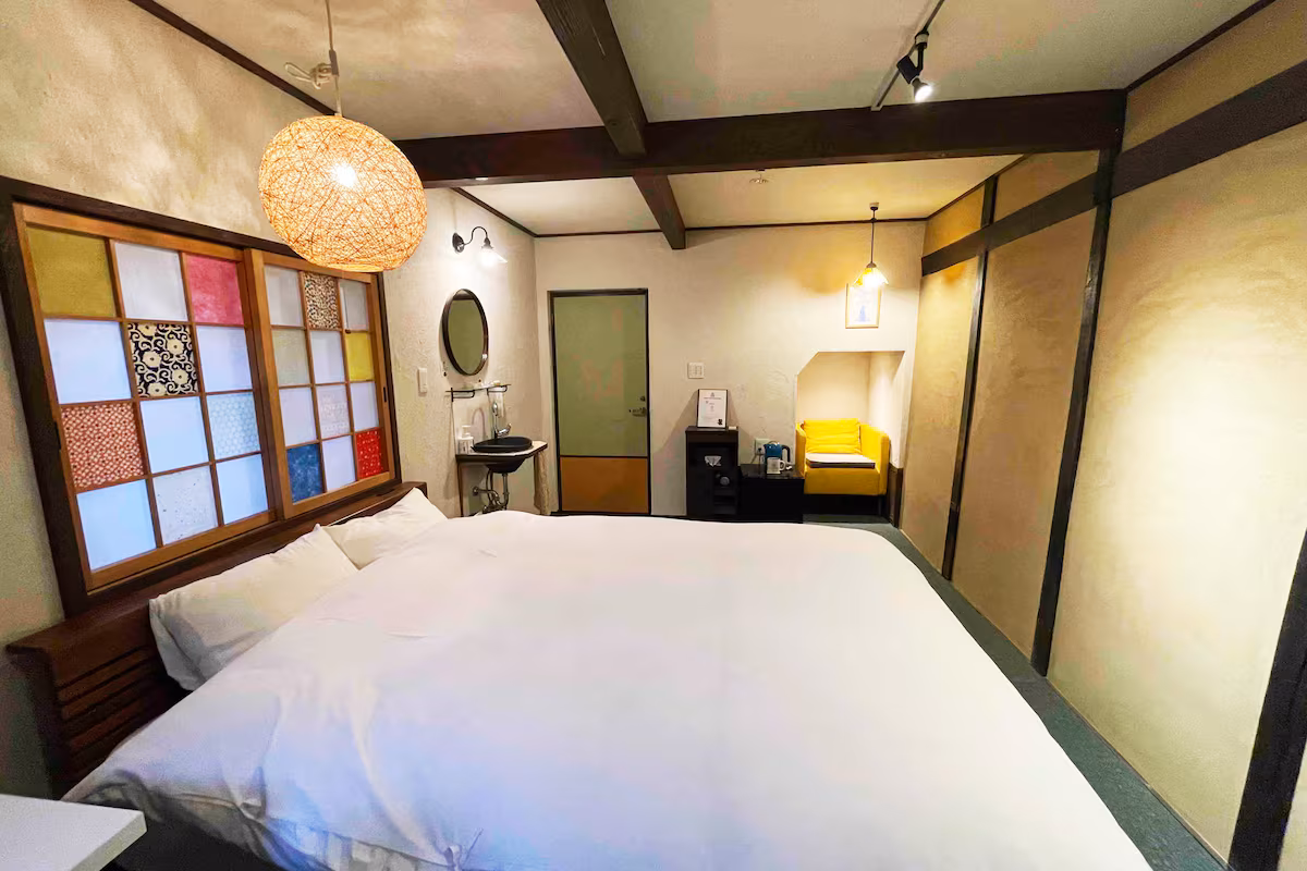 Kin room's double bed under a coloured shoji panel, with a yellow armchair and washbasin beyond