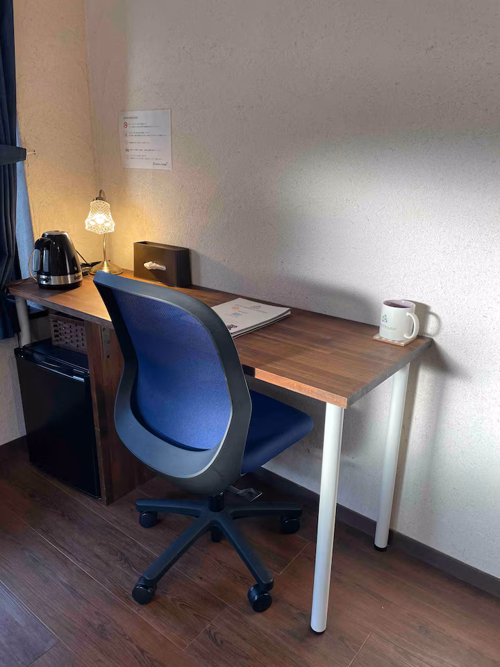 Desk in Kon room with a blue office chair, a kettle, a mug and a small fridge underneath